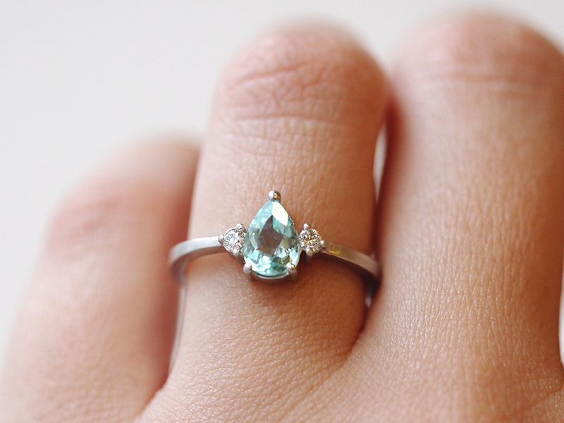 mão feminina vestindo anel de noivado em ouro 18k branco com turmalina paraíba natural em formato de gota e dois diamantes
