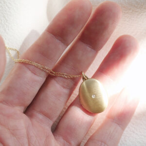 Corrente com pingente relicário em formato oval minimalista feito a mão em ouro 18k com diamante no lado de fora e dois espaços para foto em cima de mão feminina