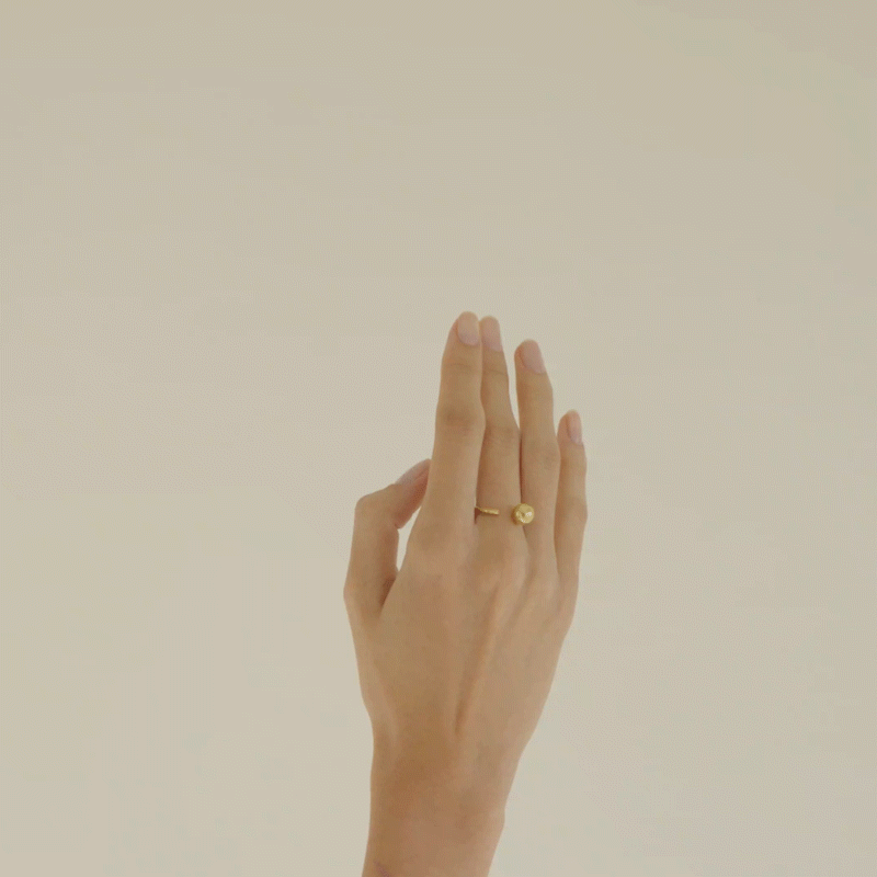 mão de mulher de pele branca usando anel em ouro amarelo 18k com detalhe de esfera se movimentando levemente em frente a fundo branco