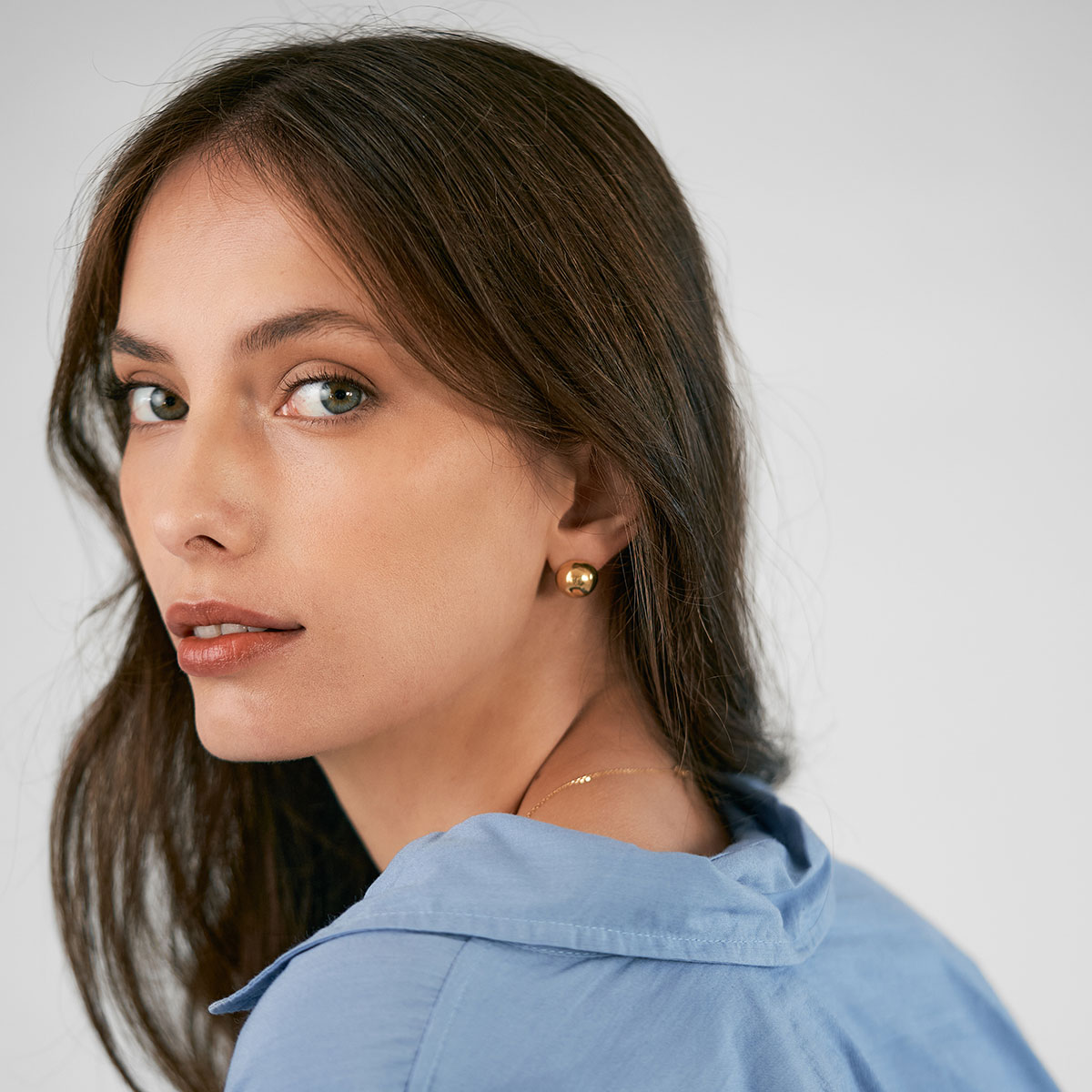mulher de cabelos longos e soltos usando camisa azul e brinco feito a mão em ouro 18k em formato de meia esfera minimalista