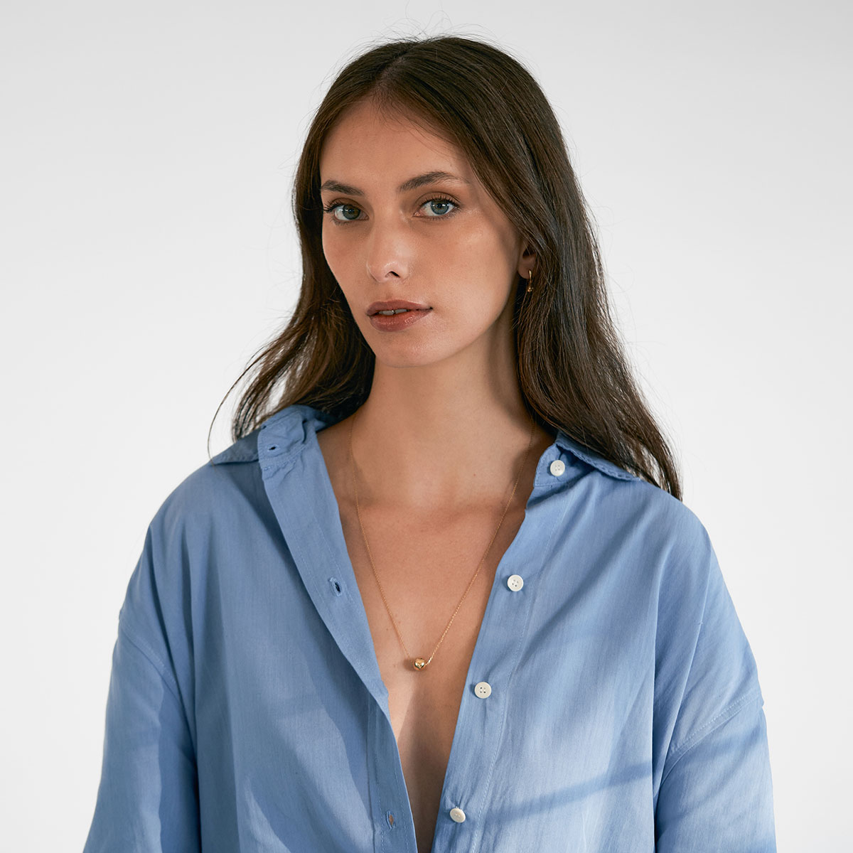 mulher de cabelos longos e soltos usando camisa azul e colar feito a mão em ouro 18k com pingente em formato de esfera minimalista