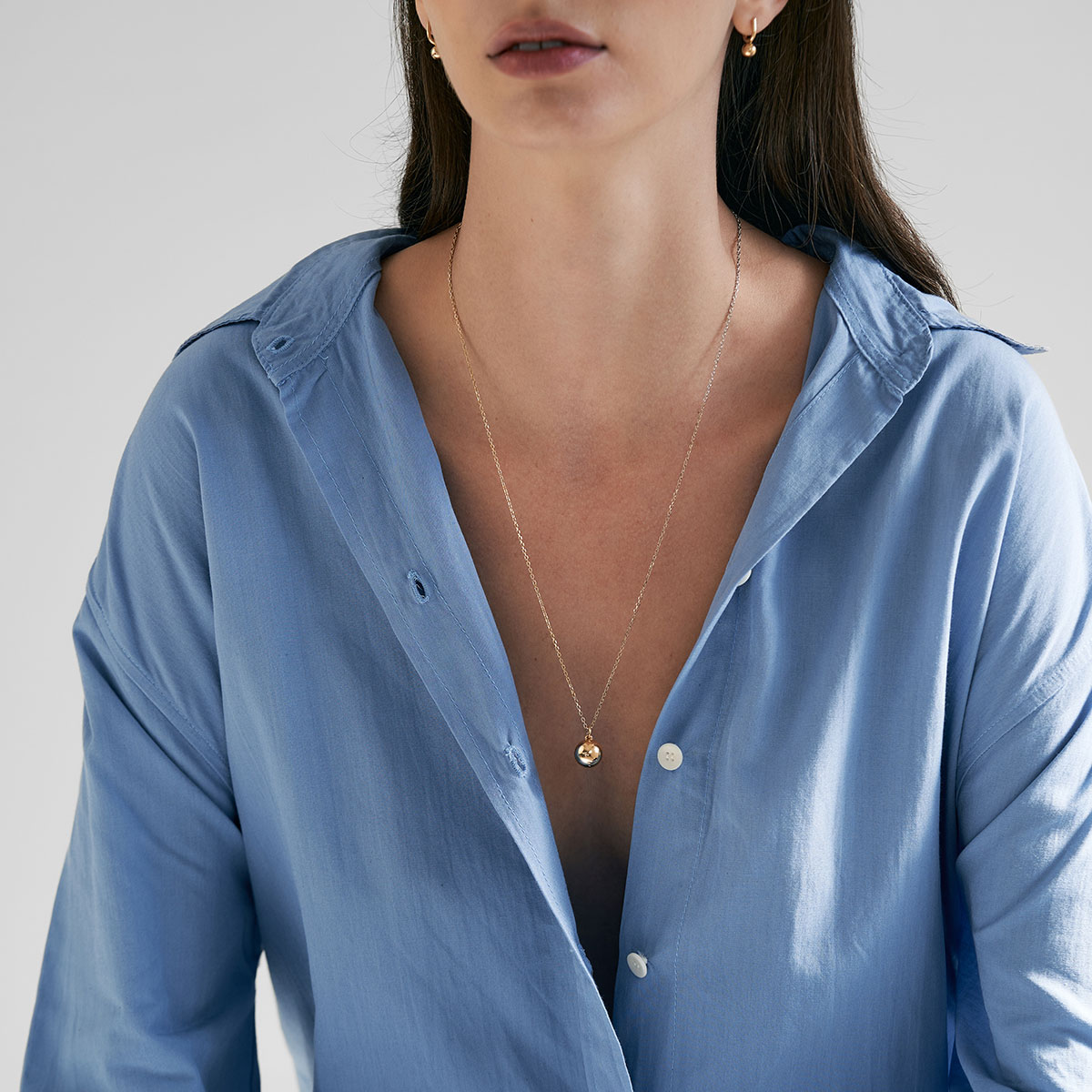 mulher de cabelos longos e soltos usando camisa azul e colar relicário minimalista feito a mão em ouro 18k para colocar fotos