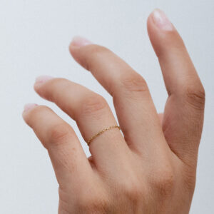 Anel de corrente em ouro 18K. Minimalista, com elos finos e singelosn anel aparece no dedo anelar de uma mão feminina.