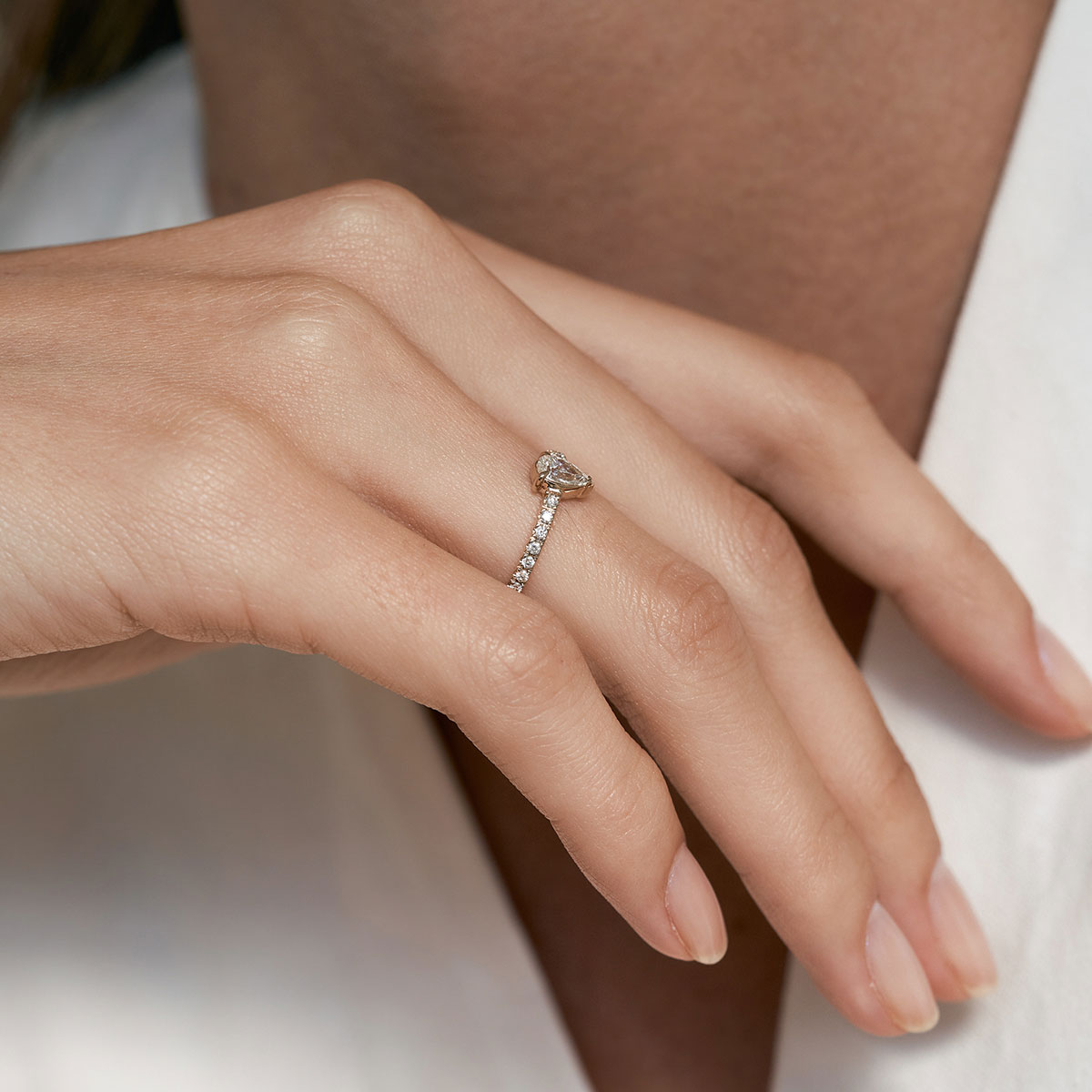 Mulher de blusa decotada branca usando no dedo anelar um Anel de noivado feito à mão em ouro 18k com aro cravejado de diamantes e no centro um diamante em formato de gota
