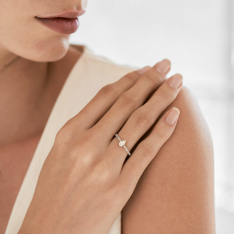 Mulher de blusa decotada branca usando no dedo anelar um Anel de noivado feito à mão em ouro 18k com aro cravejado de diamantes e no centro um diamante em formato de gota