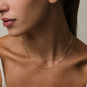 modelo loira de pele branca e cabelos presos, usando blusa bege e dois colares em ouro 18k: um com corrente tipo cartier fina e 9 pingentes com diamantes de 0,03ct e o outro uma corrente veneziana de 45cm, em frente à parede branca