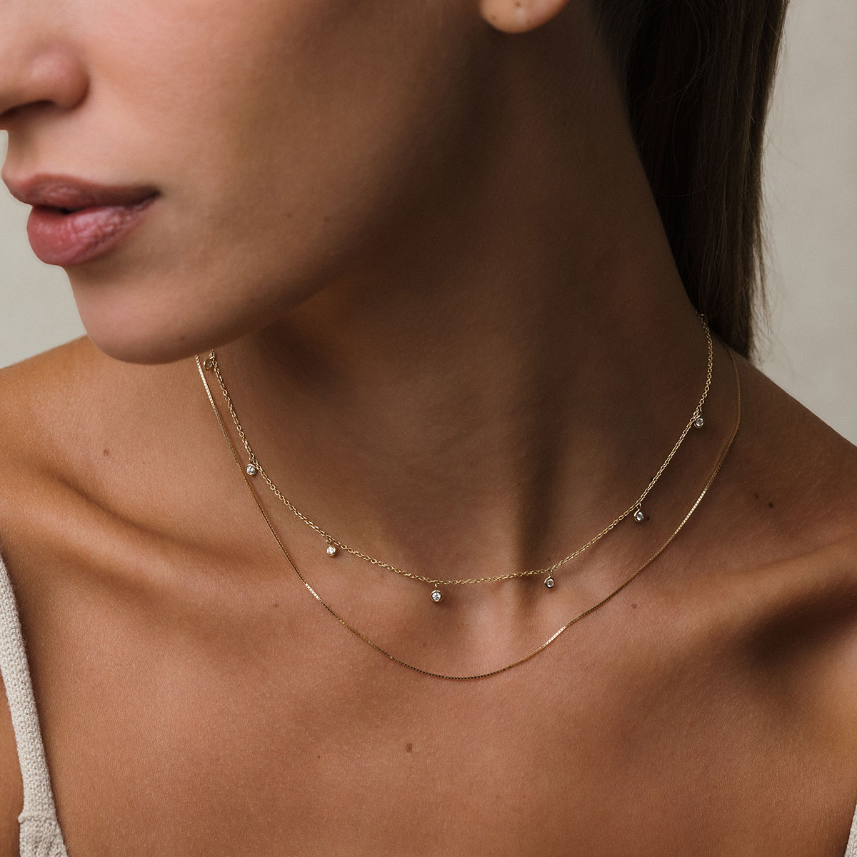 modelo loira de pele branca e cabelos presos, usando blusa bege e dois colares em ouro 18k: um com corrente tipo cartier fina e 9 pingentes com diamantes de 0,03ct e o outro uma corrente veneziana de 45cm, em frente à parede branca