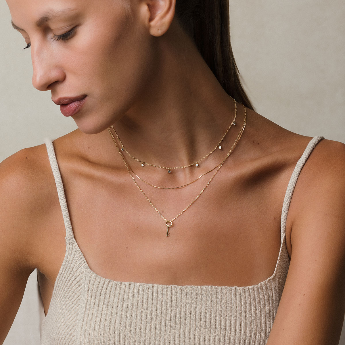 modelo loira de pele branca e cabelos presos, usando blusa bege e 3 colares em ouro 18k: um com corrente tipo cartier fina e 9 pingentes com diamantes de 0,03ct, o outro uma corrente veneziana de 45cm e o outro uma corrente cartier de elos longos com pingente em formato de chave, em frente à parede branca
