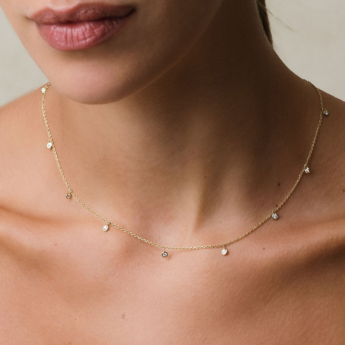 modelo loira de pele branca e cabelos presos usando colar em ouro 18k com corrente tipo cartier fina e 9 pingentes com diamantes de 0,03ct, em frente à parede branca
