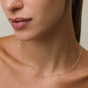 modelo loira de pele branca e cabelos presos usando colar em ouro 18k com corrente tipo cartier fina e 9 pingentes com diamantes de 0,03ct, em frente à parede branca