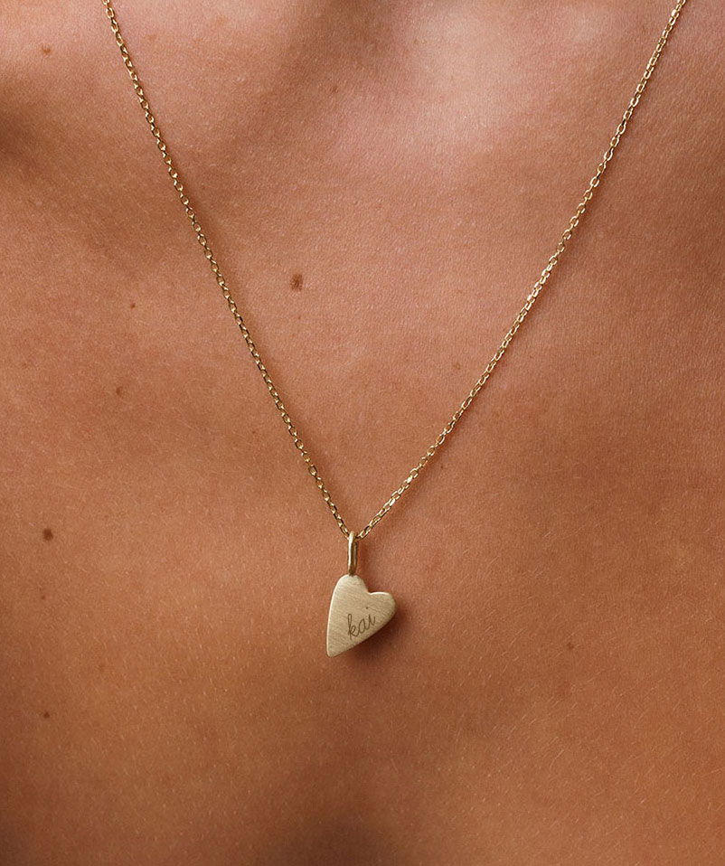 foto com foco no colo de uma mulher usando um colar e um pingente de coração com gravação escrito o nome 'kai' e com acabamento fosco em ouro 18k