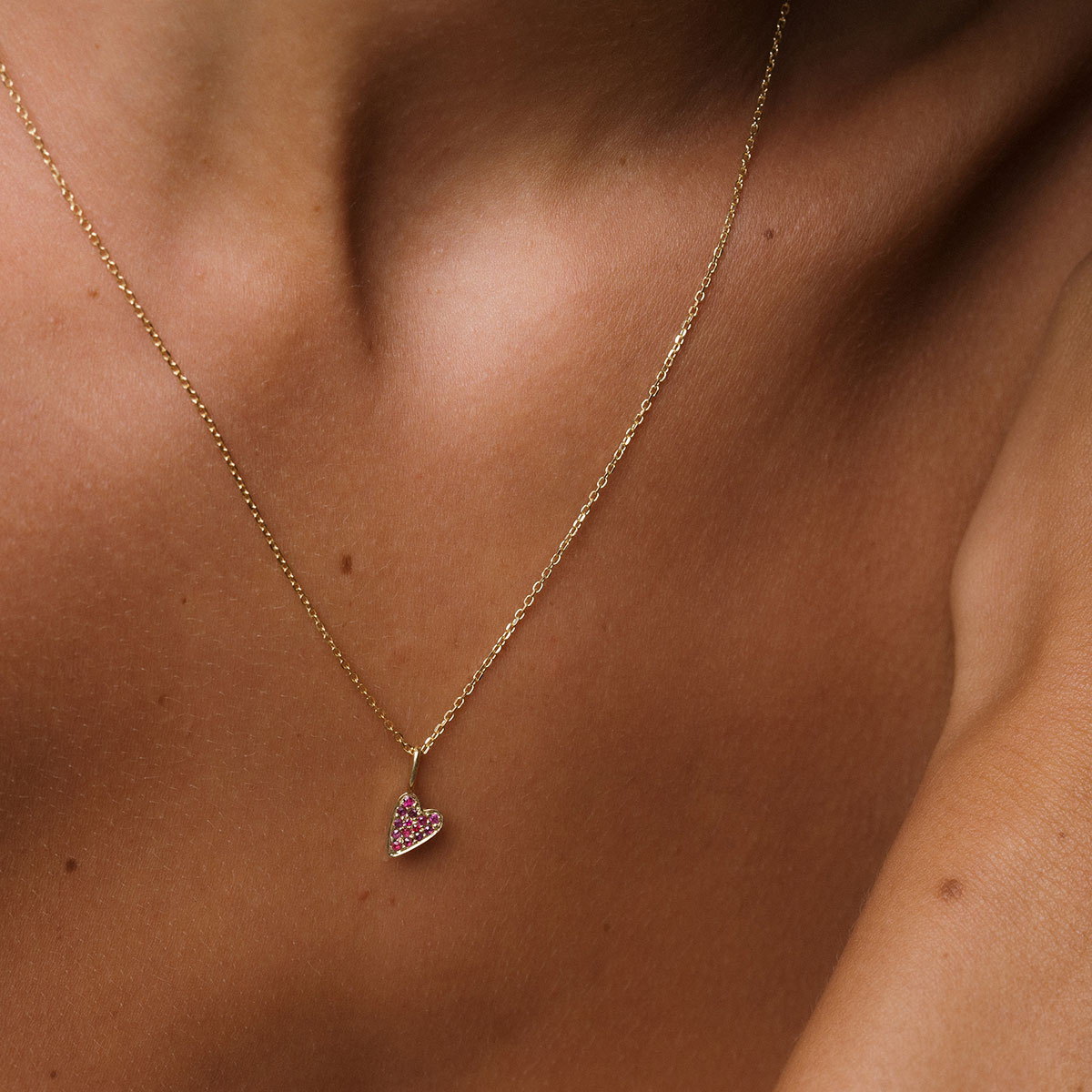 foto com foco no colo de uma mulher usando um colar de ouro 18k e um pingente de coração cravejado com 14 rubis