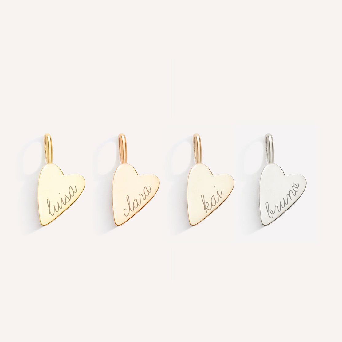 foto de quatro pingentes de coração, nas ordem de tonalidades amarelo, rosé, champagne e branco, com gravação dos nomes "luisa", "clara", "kai" e "bruno" respectivamente, em letra cursiva e com acabamento fosco, em ouro 18k, sobre fundo branco