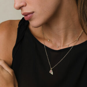 Mulher loura de cabelo presos usando blusa preta e uma colar de ouro 18k curto com nove diamantes pendurados e outro colar com dois pingentes em formato de coração, o menor deles cravejado com quatorze rubis, também em ouro 18k