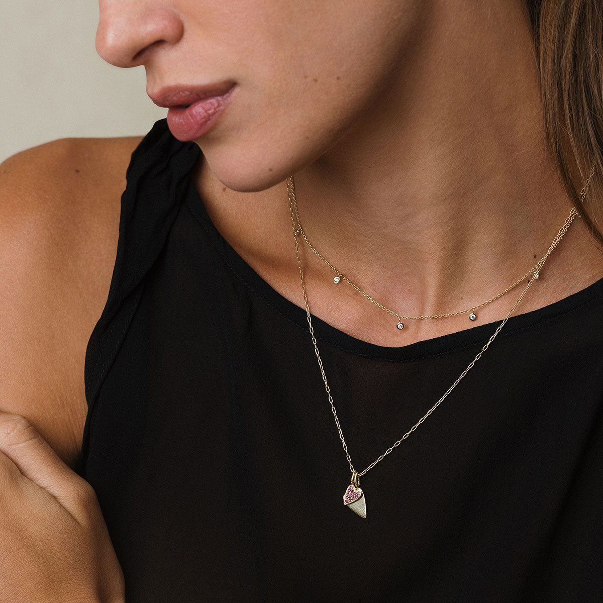 Mulher loura de cabelo presos usando blusa preta e uma colar de ouro 18k curto com nove diamantes pendurados e outro colar com dois pingentes em formato de coração, o menor deles cravejado com quatorze rubis, também em ouro 18k