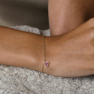 foto com foco no pulso de uma mulher usando um vestido bege e uma pulseira de ouro 18k com um pingente de coração cravejado com 14 rubis