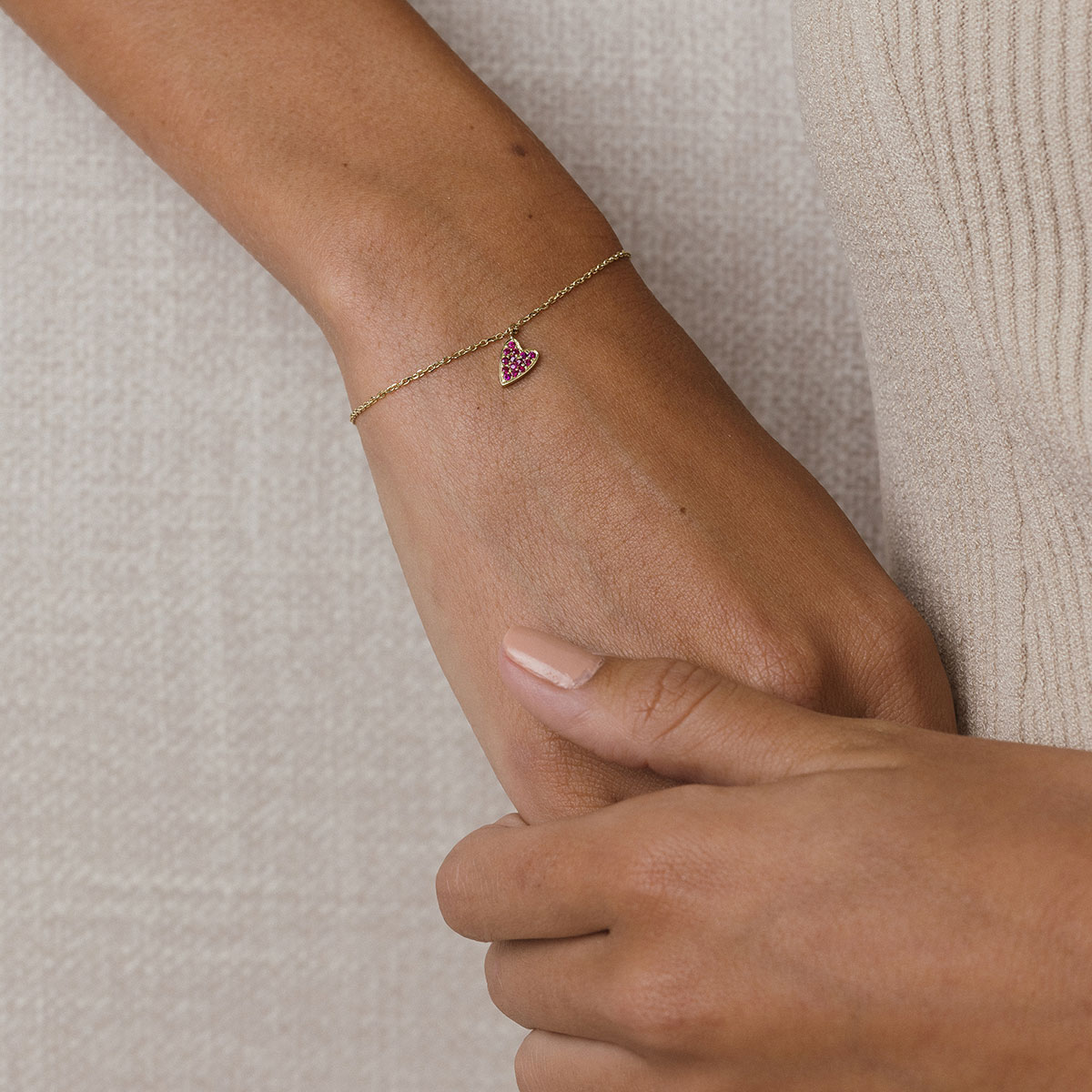 foto com foco no pulso de uma mulher usando roupa bege e uma pulseira de ouro 18k com um pingente de coração cravejado com 14 rubis