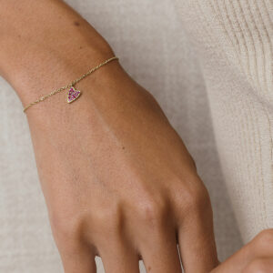 foto com foco no pulso de uma mulher usando roupa bege e uma pulseira de ouro 18k com um pingente de coração cravejado com 14 rubis