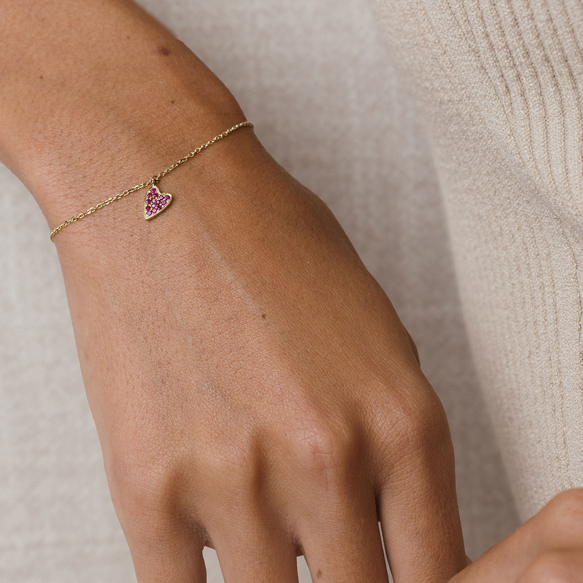 foto com foco no pulso de uma mulher usando roupa bege e uma pulseira de ouro 18k com um pingente de coração cravejado com 14 rubis