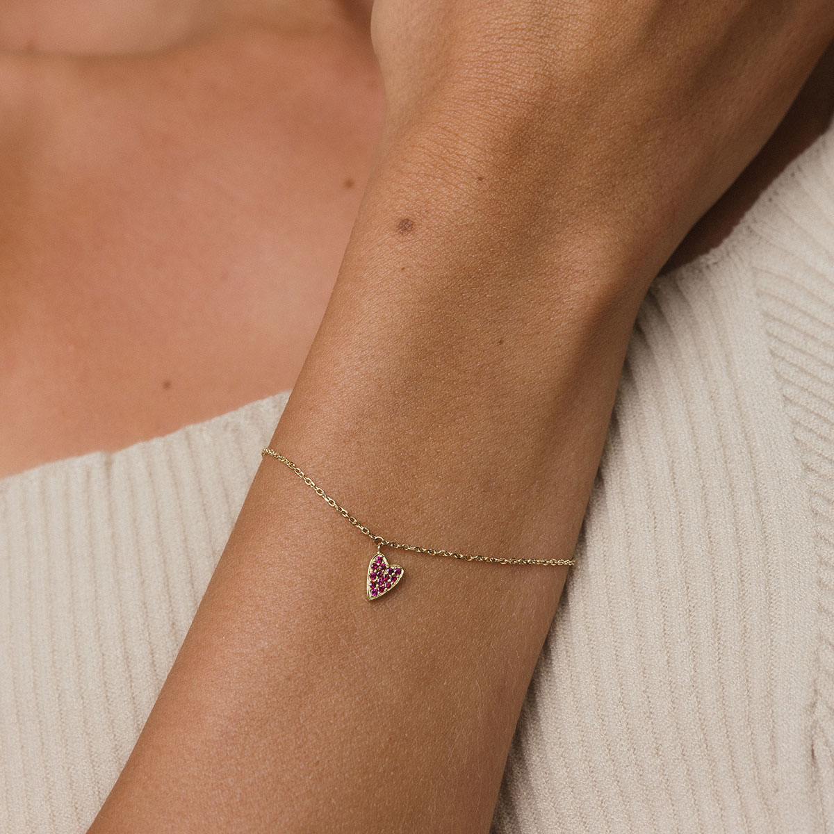 foto com foco no pulso de uma mulher usando roupa bege e uma pulseira de ouro 18k com um pingente de coração cravejado com 14 rubis