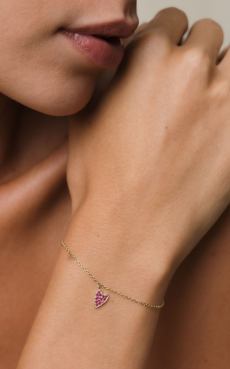 foto com foco no pulso de uma mulher usando uma pulseira de ouro 18k com um pingente de coração cravejado com 14 rubis