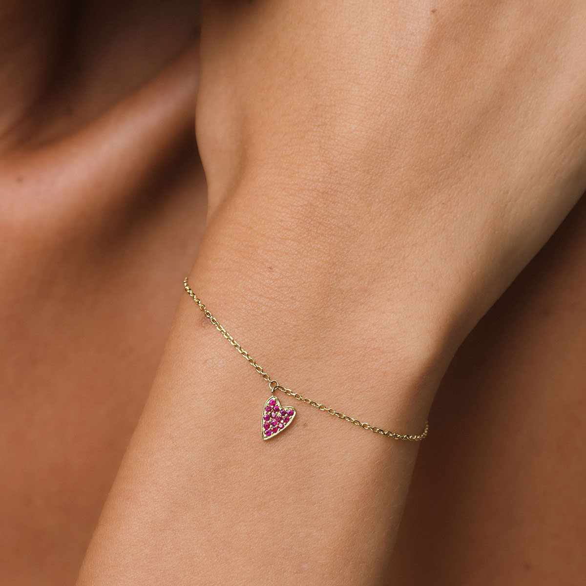 foto com foco no pulso de uma mulher usando uma pulseira de ouro 18k com um pingente de coração cravejado com 14 rubis