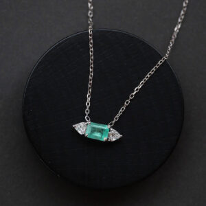 Imagem do Colar com Esmeralda e Diamantes Trillion da Joias Liê em destaque sobre fundo preto. A peça possui uma esmeralda central de 1,48 ct, em lapidação retangular, ladeada por dois diamantes com corte trillion, em uma corrente cartier de ouro branco 18k. Uma joia elegante, com proporções simétricas e brilho marcante.