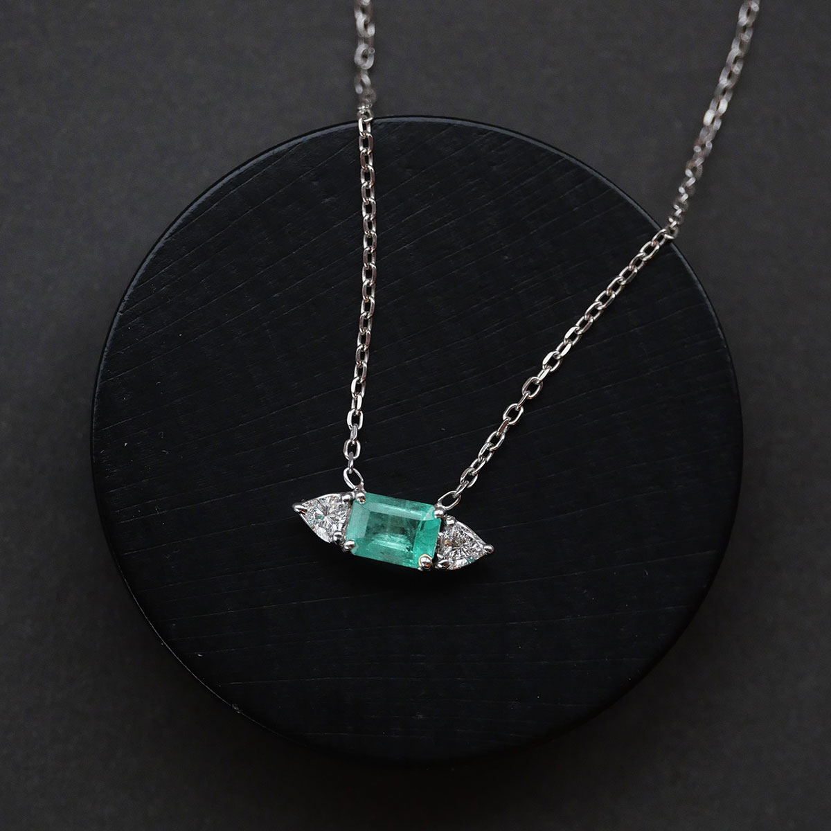 Imagem do Colar com Esmeralda e Diamantes Trillion da Joias Liê em destaque sobre fundo preto. A peça possui uma esmeralda central de 1,48 ct, em lapidação retangular, ladeada por dois diamantes com corte trillion, em uma corrente cartier de ouro branco 18k. Uma joia elegante, com proporções simétricas e brilho marcante.