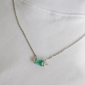 Colar com Esmeralda e Diamantes Trillion da Joias Liê em destaque sobre uma camiseta branca. A peça possui uma esmeralda central de 1,48 ct, em lapidação retangular, ladeada por dois diamantes com corte trillion, em uma corrente cartier de ouro branco 18k.