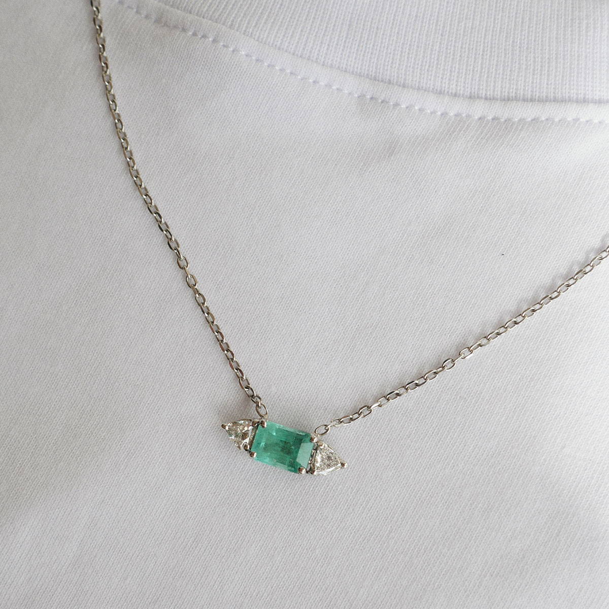Colar com Esmeralda e Diamantes Trillion da Joias Liê em destaque sobre uma camiseta branca. A peça possui uma esmeralda central de 1,48 ct, em lapidação retangular, ladeada por dois diamantes com corte trillion, em uma corrente cartier de ouro branco 18k.