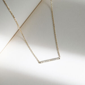 Fotografia still do Colar Traço com Diamantes da Joias Liê. A joia é feita em ouro 18k e apresenta uma barra horizontal minimalista cravejada com pequenos diamantes alinhados, conectada a uma corrente delicada. O design limpo e moderno confere leveza e sofisticação, criando um visual sutil e elegante. Ideal para uso diário ou para composições em camadas com outras peças.