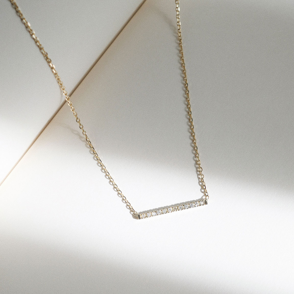 Fotografia still do Colar Traço com Diamantes da Joias Liê. A joia é feita em ouro 18k e apresenta uma barra horizontal minimalista cravejada com pequenos diamantes alinhados, conectada a uma corrente delicada. O design limpo e moderno confere leveza e sofisticação, criando um visual sutil e elegante. Ideal para uso diário ou para composições em camadas com outras peças.