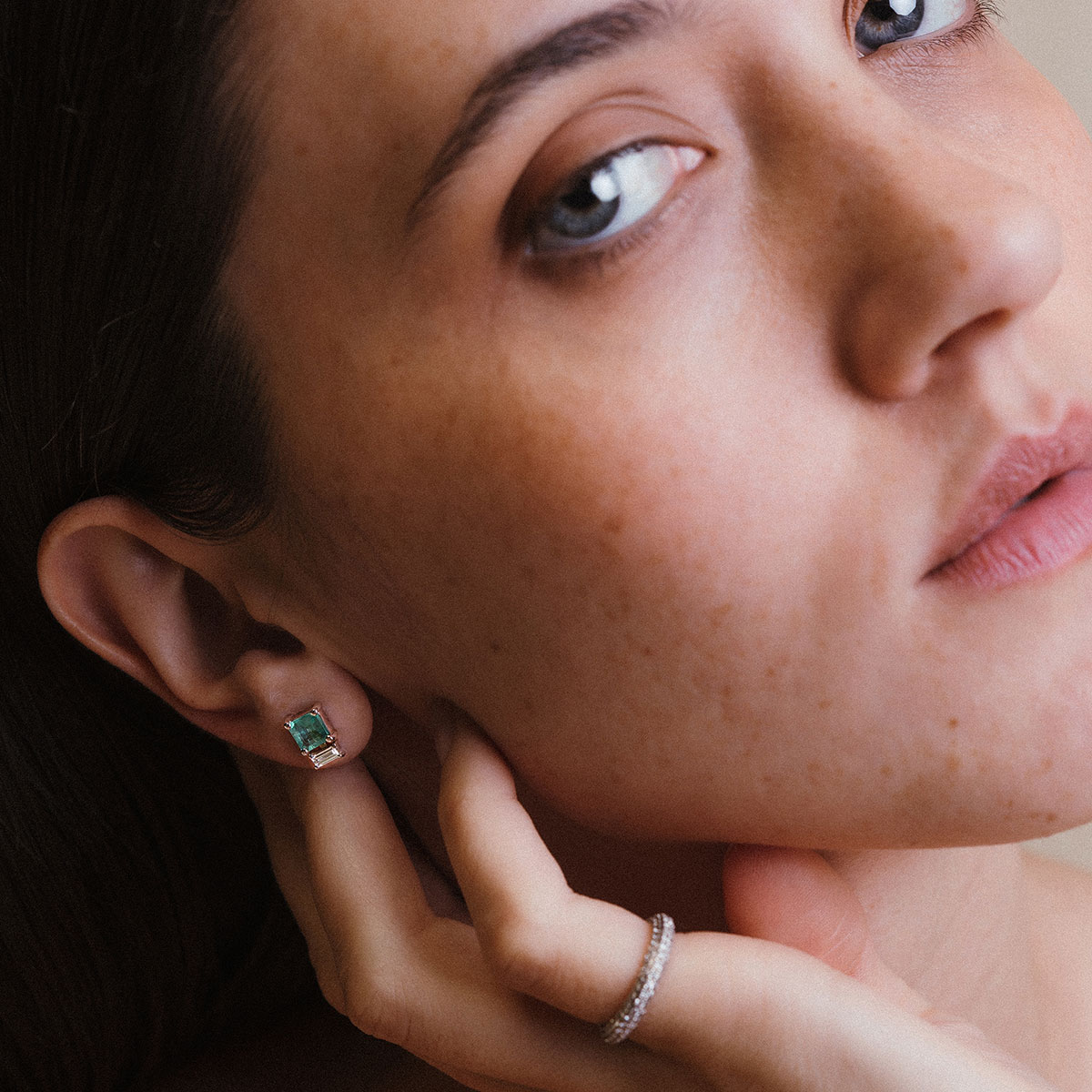 Imagem de close evidenciando o Brinco Esmeralda com Diamantes Baguete da Joias Liê. A peça apresenta uma esmeralda de 1,28 ct em lapidação retangular, ladeada por dois diamantes em corte baguete. Um design elegante, simétrico e sofisticado, que combina o verde vibrante da esmeralda com o brilho limpo e moderno dos diamantes. Também aparece o anel pinky ring bombe em ouro branco da marca
