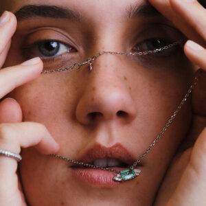 Close no rosto de mulher com colar em destaque, mostrando esmeralda central e diamantes trillion em corrente de ouro branco.