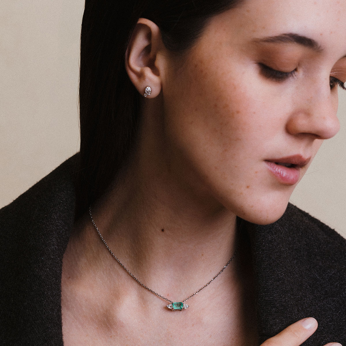 Mulher vestindo casaco preto e usando um colar com esmeralda e dois diamantes trillion da cada lado e corrente delicada em ouro branco 18k, modelo Colar com Esmeralda e Diamantes Trillion - 1,48 ct da Joias Liê
