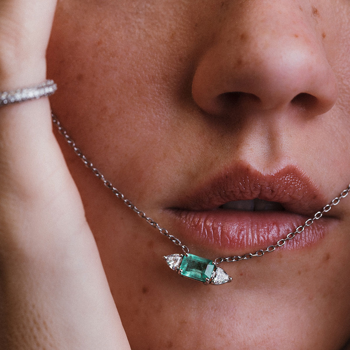 Close no rosto de mulher com colar em destaque, mostrando esmeralda central e diamantes trillion em corrente de ouro branco.
