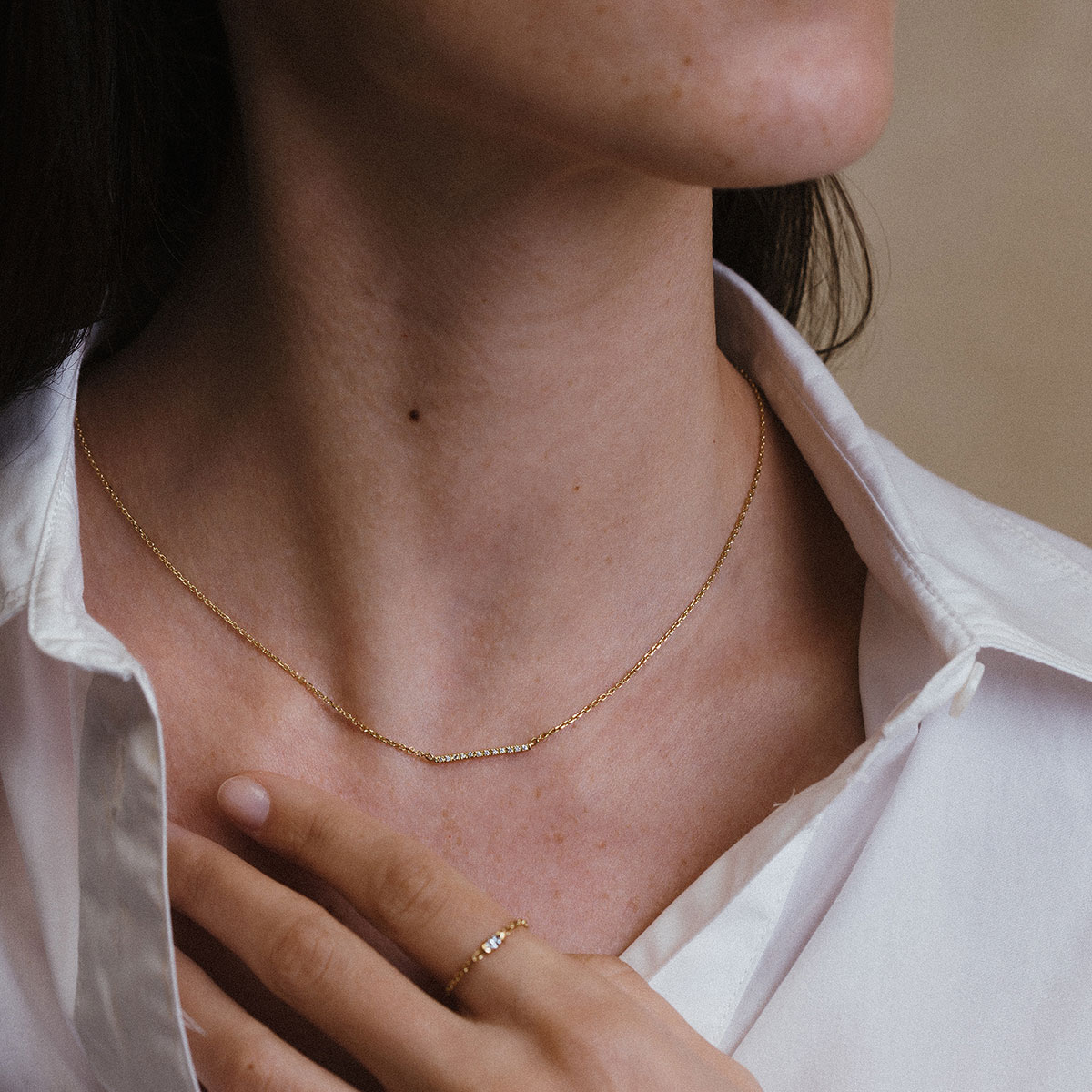 Mulher vestindo camisa branca e usando um colar com uma barra delicada e cravejada de diamantes e corrente delicada em ouro 18k, modelo Colar Traço com Diamantes da Joias Liê
