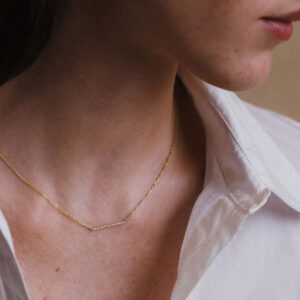 Mulher vestindo camisa branca e usando um colar com uma barra delicada e cravejada de diamantes e corrente delicada em ouro 18k, modelo Colar Traço com Diamantes da Joias Liê