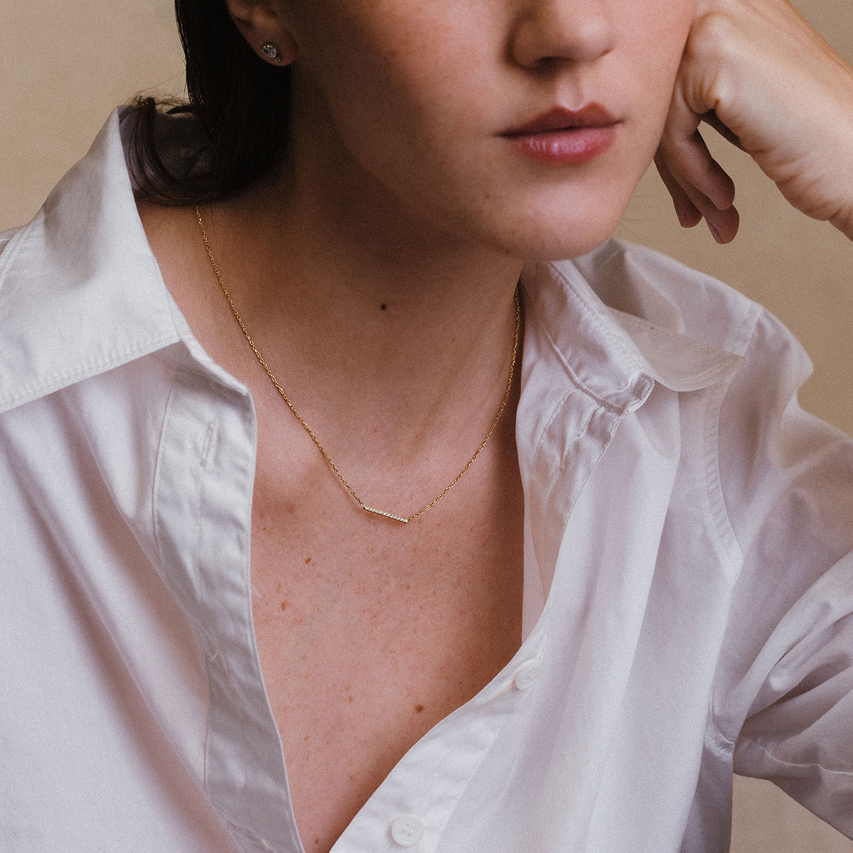 Mulher vestindo camisa branca e usando um colar com uma barra delicada e cravejada de diamantes e corrente delicada em ouro 18k, modelo Colar Traço com Diamantes da Joias Liê