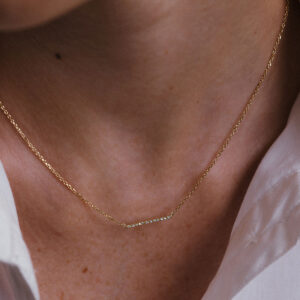 Mulher vestindo camisa branca e usando um colar com uma barra delicada e cravejada de diamantes e corrente delicada em ouro 18k, modelo Colar Traço com Diamantes da Joias Liê