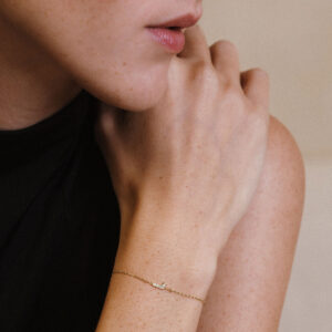 Mulher vestindo blusa preta com a mão sobre o ombro usando uma pulseira com a letra L, cravejada de diamantes lab grown, e corrente delicada em ouro 18k, modelo Pulseira Inicial com Diamantes da Joias Liê