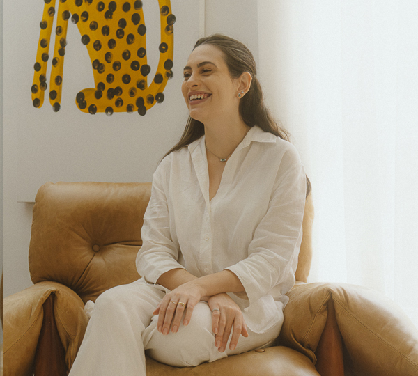 Natalia Maestrello sorri sentada em poltrona de couro, usando camisa de linho branca e joias Liê, em fotografia para a série “Em Camadas: uma série sobre propósito e autenticidade” no blog da Liê.