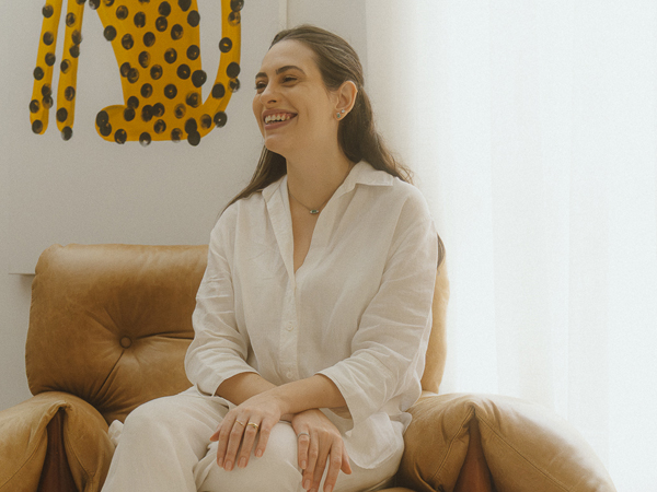 Natalia Maestrello sorri sentada em poltrona de couro, usando camisa de linho branca e joias Liê, em fotografia para a série “Em Camadas: uma série sobre propósito e autenticidade” no blog da Liê.