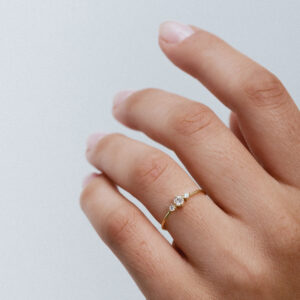 Anel em ouro 18K com três diamantes alinhados e centralizados. Anel delicado e minimalista. Aparece no dedo anelar de uma mão feminina, mais de perto mostrando sua delicadeza.