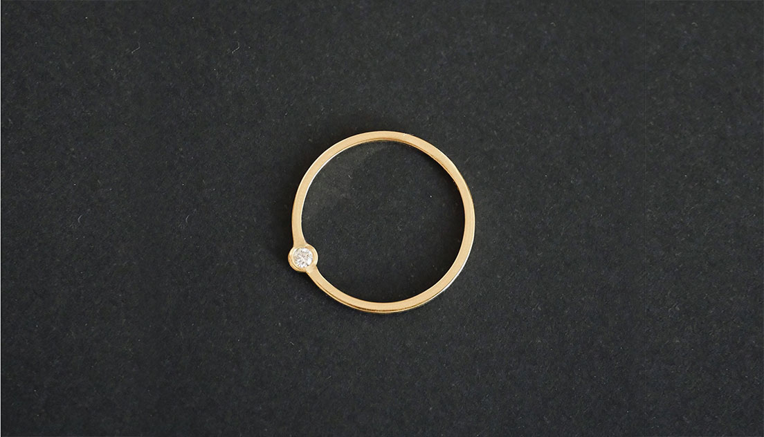 Anel Órbita em ouro 18k com diamante em design circular minimalista sobre fundo escuro.