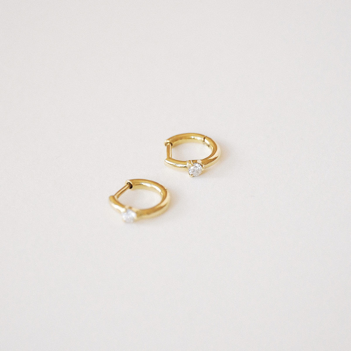 Par de Brincos Huggie Ponto em ouro amarelo 18k com diamantes sobre fundo off white.